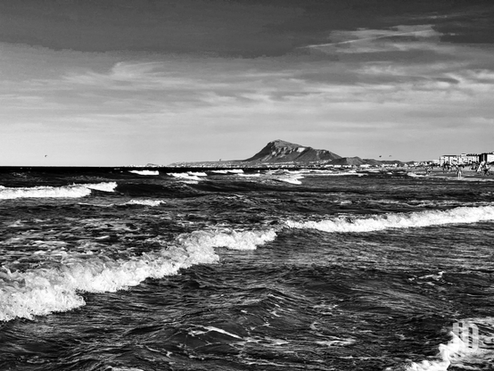 Una fotografía en blanco y negro de una escena costera. En primer plano, olas rompiendo con espuma blanca llenan el mar oscuro y agitado. A lo largo de la orilla, a la derecha, se ven siluetas de personas paseando y edificios costeros. Dominando el fondo, una gran montaña escarpada se eleva a la izquierda, con colinas más pequeñas extendiéndose hacia la derecha. El cielo está cubierto de nubes variadas, algunas más oscuras y otras más claras, creando un contraste dramático. Pequeñas cometas o parapentes son visibles en el cielo sobre el horizonte.