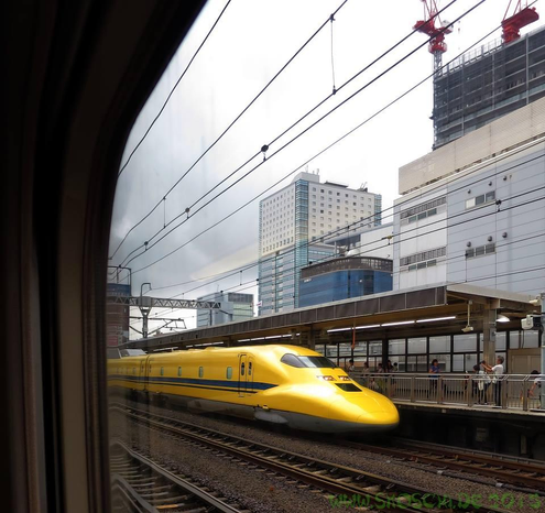 Durch ein Zugfenster beobachte ich einen gelbgefärbten Hochgeschwindigkeitszug mit lange Vorderpartie. Am Bahnsteig stehen interessierte Fahrgäste und machen Fotos. 