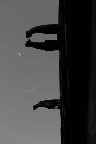 Black and white photo of three gargoyle sculptures extending from the side of a building with a crescent moon and a faint airplane contrail visible in the sky in the background.