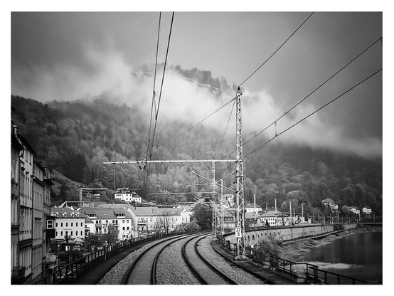 Schwarz-Weiß-Foto im Querformat mit weißem Rand. Landschaft mit Häusern links und der Elbe rechts, im Hintergrund ein Berg mit Nebelschwaden. Das Bild ist aus dem hinteren Zugfenster aufgenommen wurden, also sieht man die Schienenstränge, wie sie sich durch die Landschaft winden. 