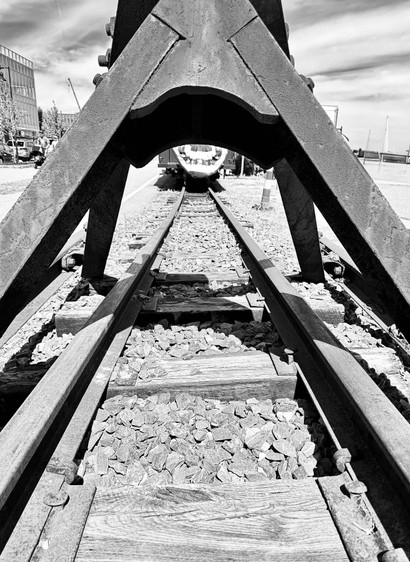 A black-and-white photo, in the foreground is a close-up of very old train tracks with horizontal wooden beams and rocks in between. Metal rails run vertically into the photo showing the perspective of distance. A bit of the back of a train can be seen with the old-fashioned round red white and blue flag on it. The rest of that train is obscured by large black metal beams coming up into a triangle shape over the tracks. The triangles on the upper right and left hand show bits of clouds in the sky and a building on the left.