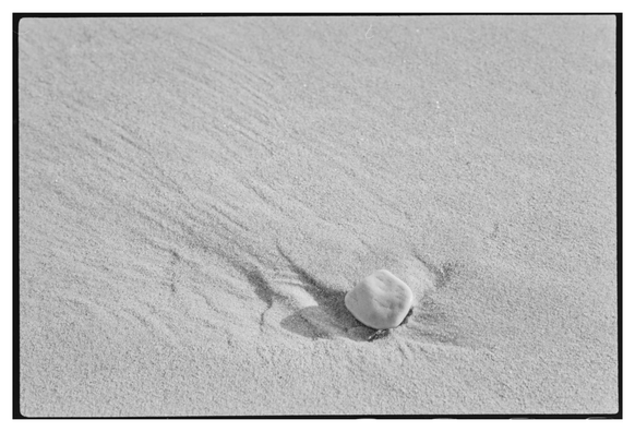 Black and white photograph in landscape orientation. High key image of a beach with a small stone. One can see in the sand that the stone has altered the flow of water around it.