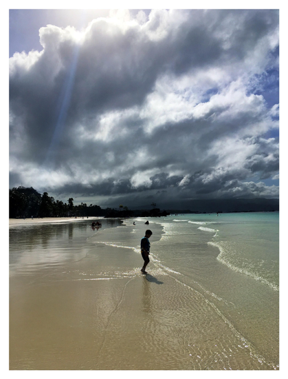 A tranquil view of the White Beach section of Boracay Island, Philippines, which has a very gradual slope allowing for a broad expanse of shallow water. Gentle waves of crystal-clear water sweep over fine, smooth sand, from the ocean at right to the far shoreline at left. A young boy stands in the middle of the photo, the water barely covering his toes. Beyond the shore are masses of trees reflected in the waves. Distant blue mountains hug the horizon. The sky is covered with cumulus clouds, dramatically backlit by the morning sun.