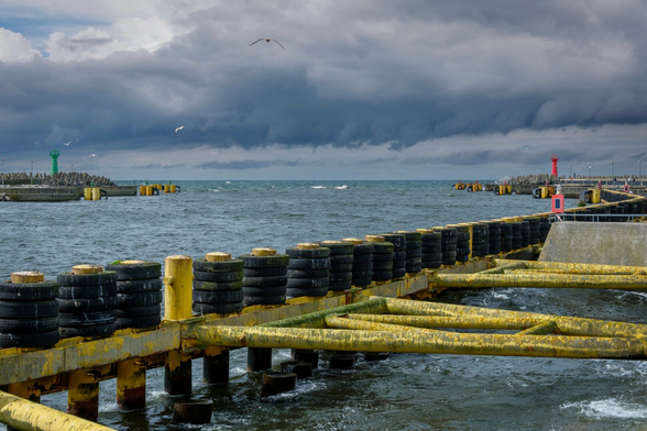 Eine Hafenszene mit einem Pier aus gelben Pömpeln und schwarzen Reifen, der eine Flussmündung entlang ins Meer unter einem bewölkten Himmel führt. In der Nähe gibt es einen grünen und roten Leuchtturm sowie einen felsigen Wellenbrecher. Möwen fliegen im rauen Wind.