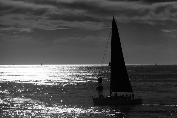 This is a black and white seascape photo of a sailing boat underway at sunset.
The lower 40% of the image is filled with an expanse of calm ocean water. 
The sea is calm with the setting sun reflecting off the surface creating large areas of silver on the left side of the image. The sea on the right is deep in shadow. To the right and below the centre of the image is a sailing boat: under sail, with the mainsail set and moving from right to left. The boat is in shadow and viewed at right angles to the camera. The crew or passengers can be seen in silhouette at the rear of the boat. A few metres beyond is a navigation buoy with a lattice tower supporting the lantern. In the distance is a crisp horizon with three further sailing boats on or close to the border between the sea and sky. Above is a partly cloudy sky with a small scattering of medium level clouds. 
The location: Redondo Beach, California (2013).