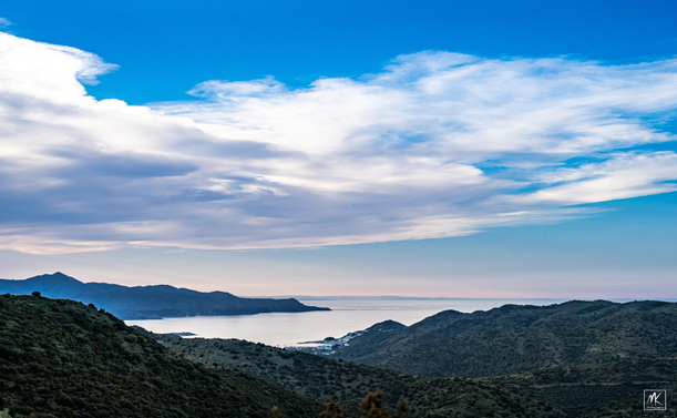 Color photo taken in the late afternoon of a mountainous coastline with clouds above. 
