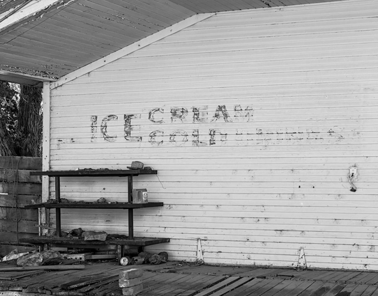 A black and white landscape photo of the left side section of a building's wood sided wall. It's an abandoned small town store. The wood plank siding is horizontal. A three tiered shelf sits on a distressed wood plank walkway on the left. A few rocks sit on the three shelves. Three bricks are stack on the wood walkway near center foreground. A brick wall with a tree trunk are seen on the far left. A sign was painted on the white wall of the building that reads, "Ice Cream Cold." There's more letters past the word cold but they are too faded to read. The legible letters are very faded as well. 