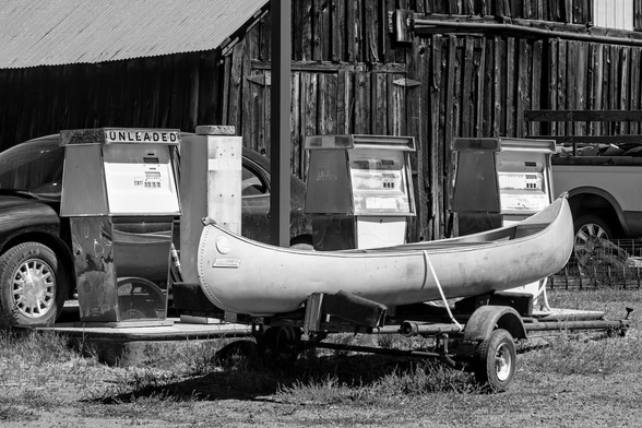 A black and white landscape photo of a aluminum canoe on a small trailer sitting in front of a line of three old, non-functioning, gas pumps. Weeds grow around the gas pump island. A black sedan is partially seen facing toward the left behind the pumps. The bed of a pickup truck is on the far right. And an old barn garage building is partially seen in the background.