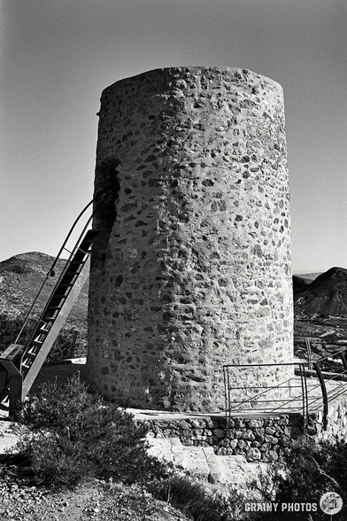 Captured with a Nikon F, this black and white photo showcases a cylindrical stone tower against a rugged, mountainous landscape. Access to an opening at a higher level is by metal steps with a handrail. Sparse vegetation clings to the structure’s base.