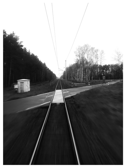 Schwarz-Weiß-Foto mit hohem Kontrast im Hochformat mit weißem Rand. Das Foto ist aus dem hinteren Zugfenster aufgenommen, daher sieht man, wie die Schienen zum Horizont hin aufeinander zuzulaufen scheinen. Die Schienen kreuzen eine Straße, dort entsteht ein grafisches Muster durch Schienen, Straße, Licht und Schatten.