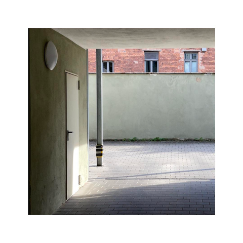 looking over a little covered parking lot into a small inner courtyard. all walls are plastered, out of concrete or red brick walls. on the left side of the frame there's a circular lamp, a door and a metal pillar that create some depth
