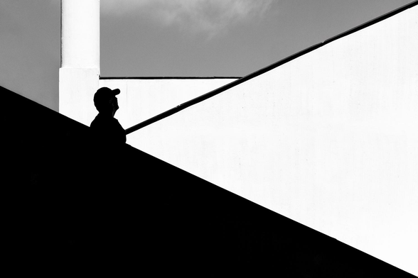The silhouette of a person stands in a stark, geometric setting. The foreground is dominated by a dark, sloping surface, contrasting sharply with the bright, minimalist background. A railing adds another linear element to the scene.