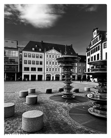 Schwarzweißfotografie: gepflasterter Marktplatz, im Vordergrund eine Springbrunnenanlage außer Betrieb, übereinander geordnete glänzende kreisrunde Scheiben, zwei solcher Stelen nebeneinander im Bild, plus zylindrische Betonsitze ringsherum verteilt, im Hintergrund alte und neue Gebäude, ein Bürgerhaus mit reicher barocker Fassade, ganz rechts Teile des alten Rathauses 