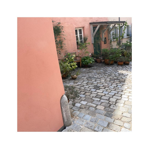 a slightly angled view at a tiny front yard built up right on the small cobblestone street. there are lots of pots and flowers all around the entrance. the surrounding walls are all painted in a subtle light pink, similar to the warm feel of brushed copper