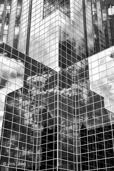 This is a black and white abstract architectural photo of a glass fronted high-rise office block.
Filling the whole image is the multi faceted corner of a high-rise office building with black reflective windows. Each horizontal surface is of a short length before turning ninety degrees inwards or outwards, making multiple corners. The surface is constructed of silver or white frames surrounding a square window. This is repeated hundreds or thousands of times across the building. Thirty floors are in view with more than that amount unseen above. In the windows is a clear view of the sky above with bright and vivid reflections of the cumulus clouds encompassing the building.
The Building: The Williams Tower, Houston (2012).