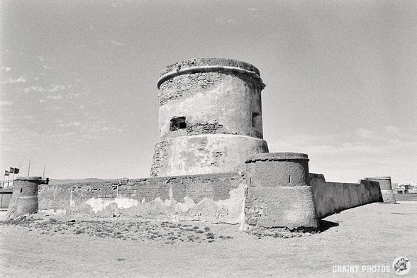 This black and white photo captures the old Tower of San Miguel, also known as the Cabo de Gata Tower. The cylindrical stone fortress is weathered and stands against a clear sky, surrounded by a low, crumbling wall. The tower is located on a beach.
