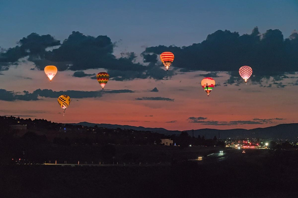 A color landscape photo of six hot air balloons with a red-orange sunrise over distant hills in the background. The six balloons are strung out from left to right at various altitudes. A line of dark clouds is behind them. All six are brightly lit and have various patterns of stripes, checkers, stars, and one has a desert scene. A highway is seen in the lower right and turning along the bottom of the photo and disappearing on the left. On the right headlights and taillights are seen on the highway. The sky is a dim blue above the clouds and balloons.