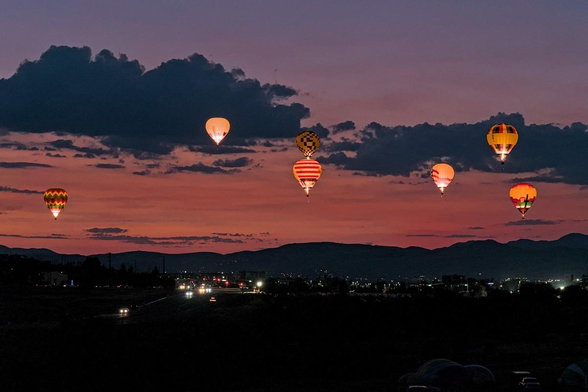 A color landscape photo of hot air balloons floating in a sunrise colored sky above a distant mountain range. There's a layer of clouds running across the sky a little above center. The sky is a light blue-red above the clouds. Below the clouds it is bright red-orange. There's a bit of a highway seen just below center left with city lights seen on either side. The seven balloons are all brightly lit. They have various geometric patterns of stars, stripes, and checkers in many colors. One has a red mesa desert scene. They are scattered across the frame at various elevations. 