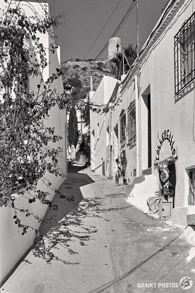 A narrow, winding alley in Nijar with whitewashed buildings and wrought iron window bars unfolds like a fairytale scene. A vine with leaves and shadows falls over the path, and an intimate shop is on the right. On the hilltop above a tower is visible.