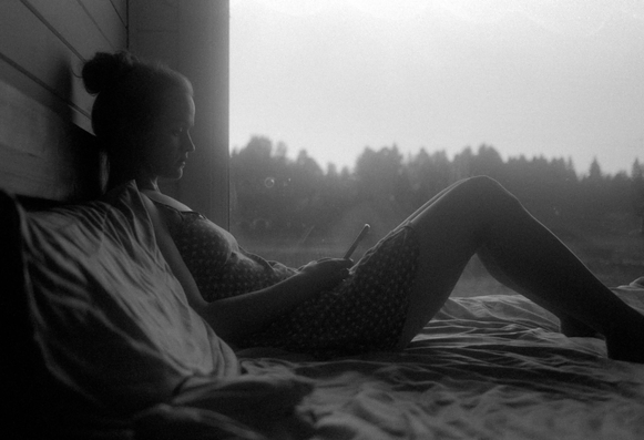 The black and white photo shows a woman sitting on a bed with a pillow under her back in a room with a view of nature. She is wearing a dress with a small pattern, and she is holding a phone in her hands. She is reading something. In the background, a foggy landscape with trees can be seen through a large window.