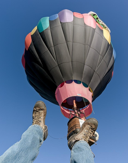 A color portrait photo of a hot air balloon from underneath. In the lower left foreground a pair of jean clad legs with hiking boots on are seen as if the photographer had been thrown out of the rising balloon and was taking a picture as he fell away from the balloon's passenger basket. The balloon is mostly black with some colored dots barely visible alone the top. A flame can be seen as the balloon accelerates away.