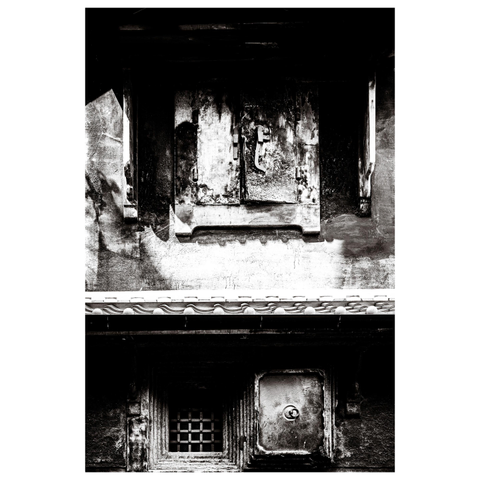 A black-and-white photo showing two sections of a weathered Japanese building façade. The upper part has a thick, decaying wooden shutter with visible cracks and peeling surfaces. Below, a smaller barred window sits next to a square iron shutter with a circular handle. The textures of rust, soot, and erosion dominate the scene, giving the impression that these shutters have withstood centuries of weather, fire, and possibly disinterest.