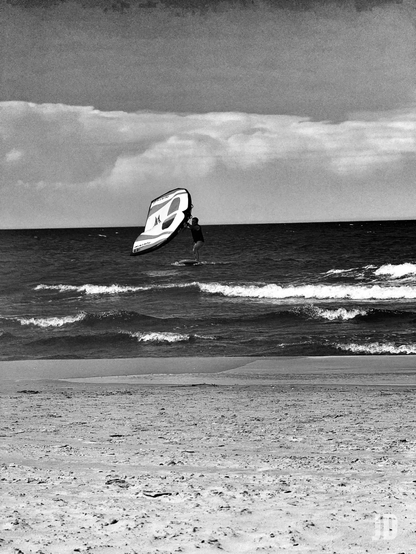 En la imagen en blanco y negro, un hombre practica wing foil en la orilla del mar. Él se encuentra de pie sobre una tabla de surf con un hidroala, mientras sostiene una cometa inflable o ala de mano que lo impulsa. Las olas rompen cerca de la playa, y el cielo está parcialmente cubierto de nubes.