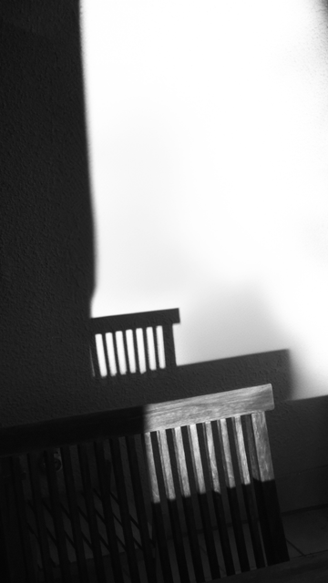 Die Rückenlehne einer Sitzbank aus Holz, die oben aus einem querlaufenden Fries und unten aus senkrechten Stäben besteht, wirft durch Sonneneinstrahlung ihren Schatten nach hinten auf eine weiße Wand. Dabei wird nicht die gesamte Rückenlehne von der Sonne angestrahlt: Der linke Teil von ihr sowie der linke Teil der weißen Wand hinter ihr liegen im Schatten.