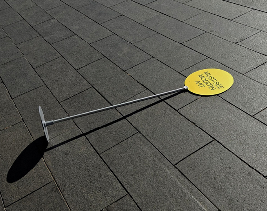 A photo of a yellow circular sign reading “MUST-SEE MODERN ART” attached to a metal pole, which has fallen over and lies flat on a tiled pavement.

