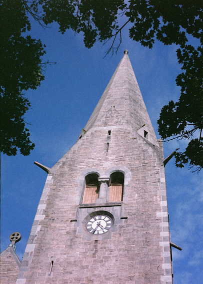 Analog photograph of a church tower built of rough stone bricks. The tower is fairly plain, with a small clock and two door-shaped windows high up on the wall where the bells probably are, but the windows are shut with wooden boards. The tower sits in pale afternoon sun against a blue sky, framed by the shady crowns of nearby trees. The colours are all a little off, due to the expired film stock.