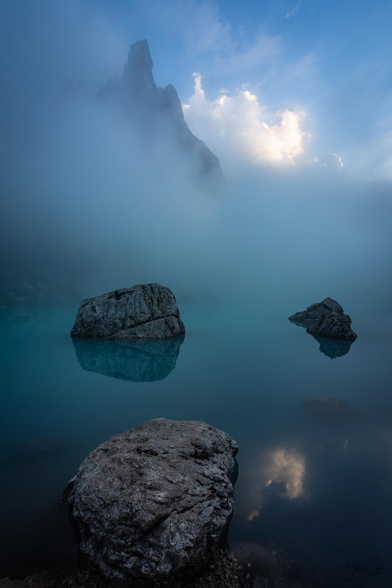 Three round rocks emerging from a turquoise blue foggy mountain lake with a rocky mountain peak appearing in the fog in the background in the left corner. In the right corner, the blue sky bathed in the late afternoon light and underlined with a puffy white cloud. All elements are relected in the lake.