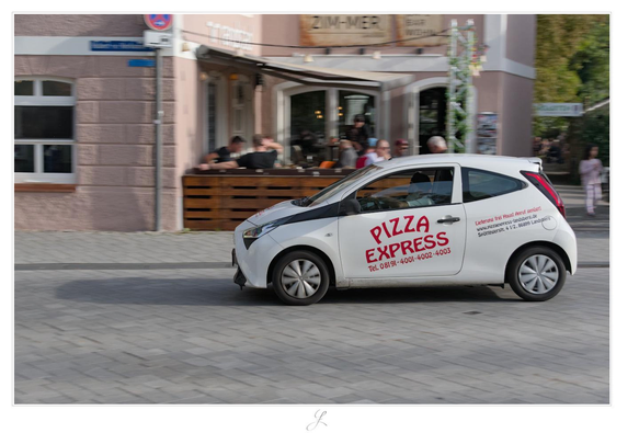 A photo of a passing car in landscape format. It is a fairly small, white car. The word "PIZZA" is written in large, slightly curved red letters on the driver's door. Below that is written "EXPRESS" and below that a phone number. Behind the driver's door is a smaller block of text with the red inscription "Lieferung frei Haus! Anruf genügt!" ("Free delivery! Just give us a call!"). Below that, an internet address and a postal address are written in black letters. The car is moving, and the camera was pulled along with it, causing the background to be blurred. The scene is rendered in warm colors, as the photo was taken on a late sunny afternoon. 

AI disclaimer: Using my work, its meta data, written or derived description to create media with or train AI based systems is prohibited.