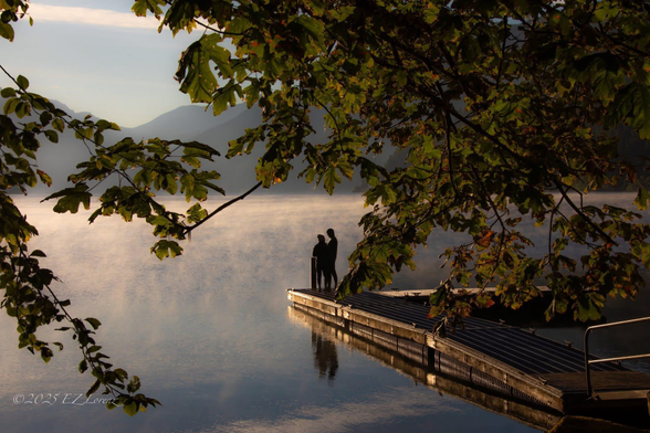 This image captures a serene moment at sunrise on Lake Crescent in Olympic National Park, Washington. 
The photo, titled "Intimate moments at sunrise," was taken on August 21, 2025 by EZ Lorenz. 
It features two figures silhouetted on a dock, gazing out over the misty lake, framed by tree branches. 