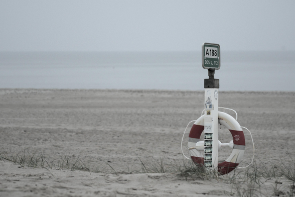 Ein Rettungsring in rot-weißer Farbe hängt an einem Pfosten mit der Nummer 'A188'. Der Hintergrund zeigt einen ruhigen Strand und das Meer.