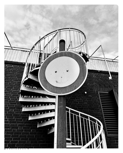Schwarzweißfotografie: eine weiße Wendeltreppe aus Metall an einem Gebäude, davor ein rundes Verkehrszeichen „Durchfahrt verboten“, dieses ist mittig mit zwei Muttern verschraubt, diese fungieren als Augen, jemand hat mit dünnen schwarzen Linien eine Nase als runden Punkt und ein lächelnden Bogen als Mund dazu gemalt 