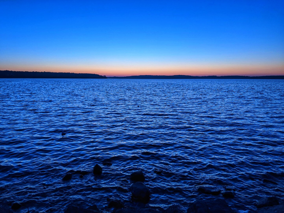 A horizontal, wide-angle shot of a lake at dusk. The sky is a deep, vibrant blue at the top, fading to a lighter blue and then to a soft orange and yellow at the horizon. The horizon is marked by a dark silhouette of a distant forest. The lake's surface is a rich, dark blue with small ripples reflecting the last light of the day. A few small, dark rocks are visible in the foreground, partially submerged in the water. The overall tone is calm and serene.