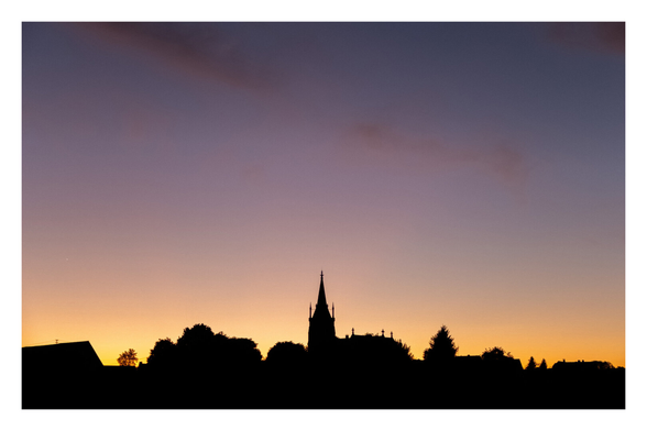 Foto im Querformat. Kurz nach Sonnenuntergang. Unten ist die schwarze Silhouette eines Stadt. Bäume, Häuser, mittig eine Kirche. Darüber ist ein Streifen orange-rötlicher Himmel, darüber ist er blau. Von links oben kommt ein dünner Streifen Wolken ins Bild.