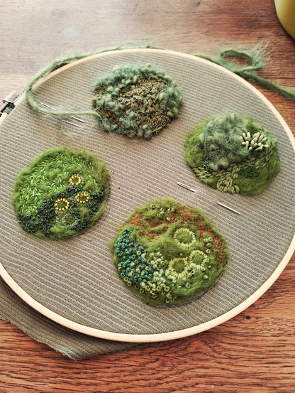 An embroidery hoop holding four textured moss embroideries in different shades of green. Each circular patch is densely stitched and layered to resemble natural mossy mounds, with varied stitches and bead details. A needle and loose green thread rest nearby.