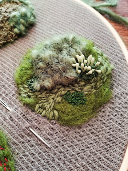 Close-up of one moss embroidery in the hoop. The patch is a mix of soft green wool, textured stitches, and tiny details, resembling a tuft of moss with small white mushroom-like shapes growing from it.