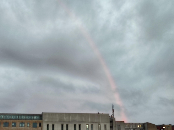 Graue Wolken mit leichtem rosa Schimmer über Hausdächern, rechts im Bild ein Regenbogen, auch er wirkt nur rosa. 