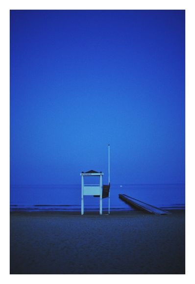 A portrait orientation photograph shows a beach at twilight. A white lifeguard tower stands in the center foreground, and a wooden pier is visible to its right. The calm sea stretches across the horizon, meeting a clear, deep blue sky.