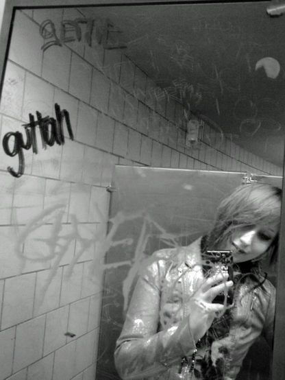 A black and white selfie in a dirty public restroom mirror, reflecting a ginger gremlin wearing a shiny jacket and holding a phone. The mirror is covered in graffiti and grime of questionable origin.