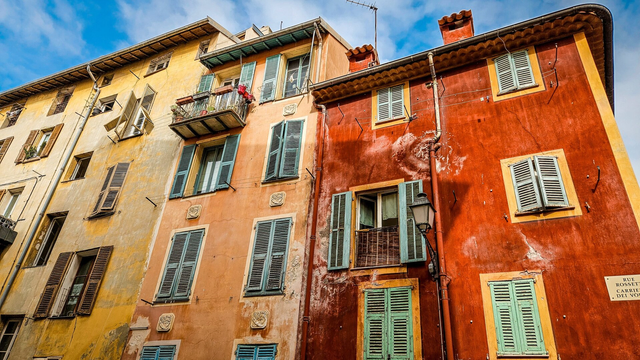 Colorful old European buildings with weathered facades in shades of yellow, peach, and deep red. The walls are lined with wooden shutters painted in muted greens and browns, some open and some closed. One balcony has flower pots, and a vintage streetlamp is attached to the red wall. The sky above is bright with scattered clouds.