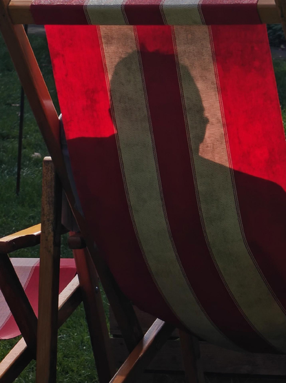 A photo of the back of a red and beige deck chair. Warm sun light casts a shadow of a person’s head and shoulders on the fabric of the chair.

