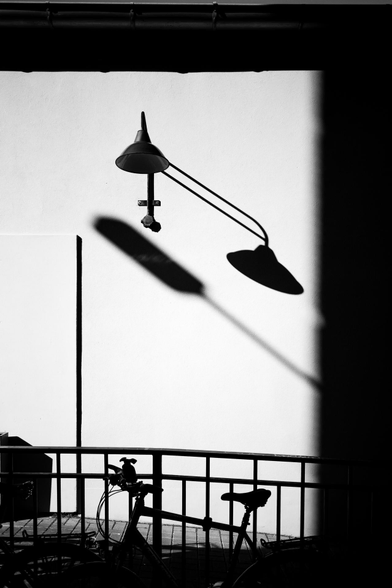 A black and white image of a minimalist urban scene. 


A simple, wall-mounted lamp casts a long, dramatic shadow across a light-gray wall. The lamp's shape and the way the light falls create a strong graphic element. 


A bicycle is silhouetted against a metal railing in the foreground, adding a sense of scale and presence to the otherwise empty space. The railing and the bicycle are in sharp contrast to the smooth, unbroken texture of the wall. 


The overall effect is one of quietude and stark contrast, with the simplicity highlighting the interplay of light and shadow.