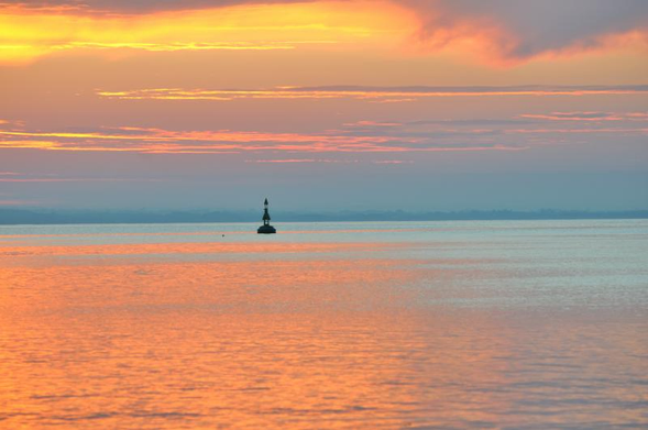 Une photo en couleur d'un coucher de soleil sur la mer. L'horizon est à peu près au tiers supérieur de l'image, le ciel offre une très riche palette qui va du jaune au mauve en passant par l'orange et le bleu voilé. Une bouée de balisage , tout près du milieu de l'image, capte le regard sur une mer ou l'orange  l'emporte sur un clair vert émeraude.
