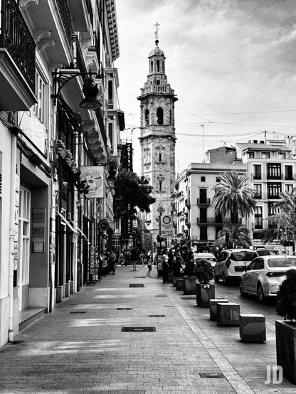 Fotografía en blanco y negro de una calle urbana que se extiende hacia una alta torre campanario al fondo. A la izquierda, una fila de edificios de estilo clásico con balcones de hierro forjado y tiendas en la planta baja, con carteles y farolas colgantes. La acera, de adoquines, está bordeada por maceteros cuadrados de piedra. A la derecha, en la calzada, hay coches estacionados y en movimiento, y se observan varias personas caminando por la acera y la calle. Más allá de los coches, se aprecian otros edificios urbanos y palmeras. La torre campanario, de piedra clara, es alta y ornamentada, con varios niveles de arcos y un chapitel en la cima. El cielo está cubierto de nubes, lo que crea una luz difusa que realza los contrastes de la imagen en blanco y negro.