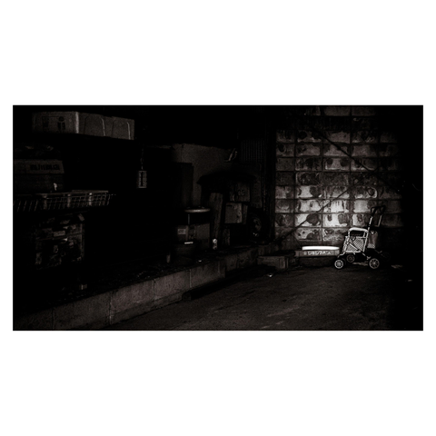 A black-and-white photo of a dimly lit storage-like space. Against the right wall, a four-wheeled walker with a patterned seat is illuminated by a narrow beam of light. The rest of the scene—boxes, shelves, and a stained concrete wall—fades into darkness.