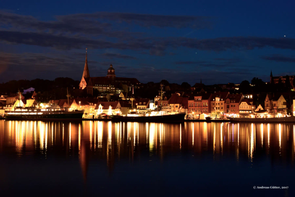 Einige alte Dampfer liegt am Kai der Flensburger Förde. Es ist Abenddämmerung, und die Stadt leuchtet golden über die Förde. Die Lichter der Stadt und der Dampfschiffe spiegeln sich und glitzern im dunklen, ruhigen Wasser der Ostsee. 