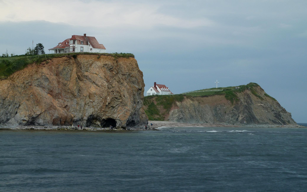Photograph of a coastal scene featuring two houses perched on brown cliffs, with rocky formations and a beach below. On the left is a large cliff with a white house with red roof at the top, and on the right another similar house further back on a grassy area, with a white cross near the edge of the cliff. The scene includes waves lapping at the shore, and a rocky beach with a couple of people. The sky is overcast.